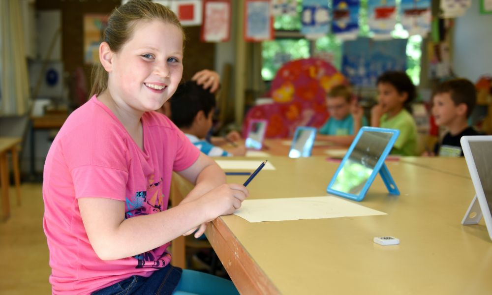 Smiling young girl in a pink shirt sitting at a desk with a piece of paper and tablet 